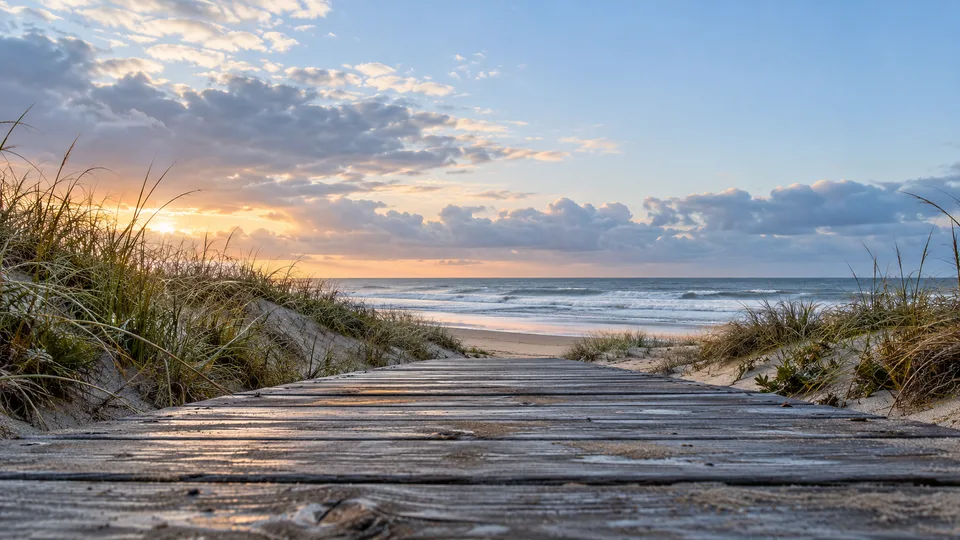 A wooden boardwalk runs between coastal dunes toward a quiet beach after rain.