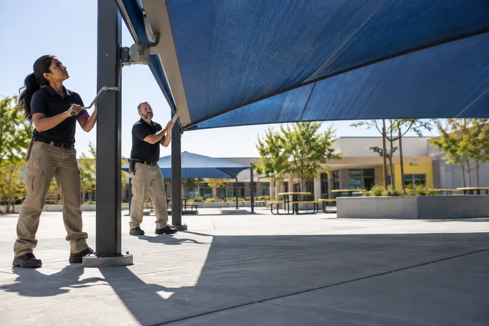 Two adults inspect an anchored blue canopy over an empty school play area on a sunny day.