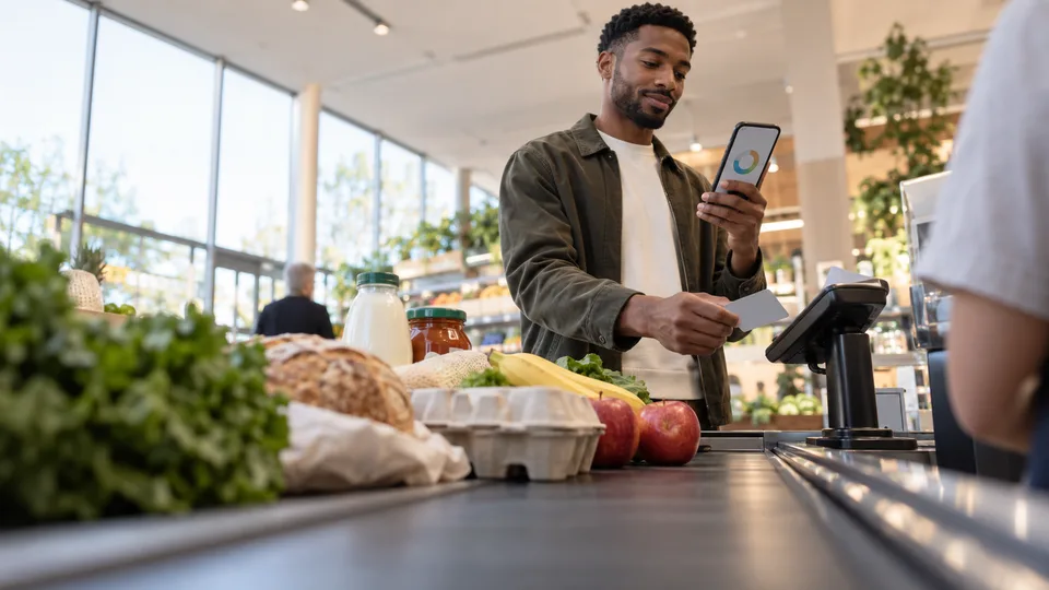 A shopper pauses beside groceries and a plain payment terminal at a checkout counter.