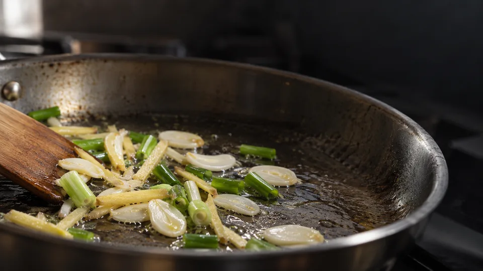 Garlic, ginger and scallions begin to saute in a pan under angled kitchen light.