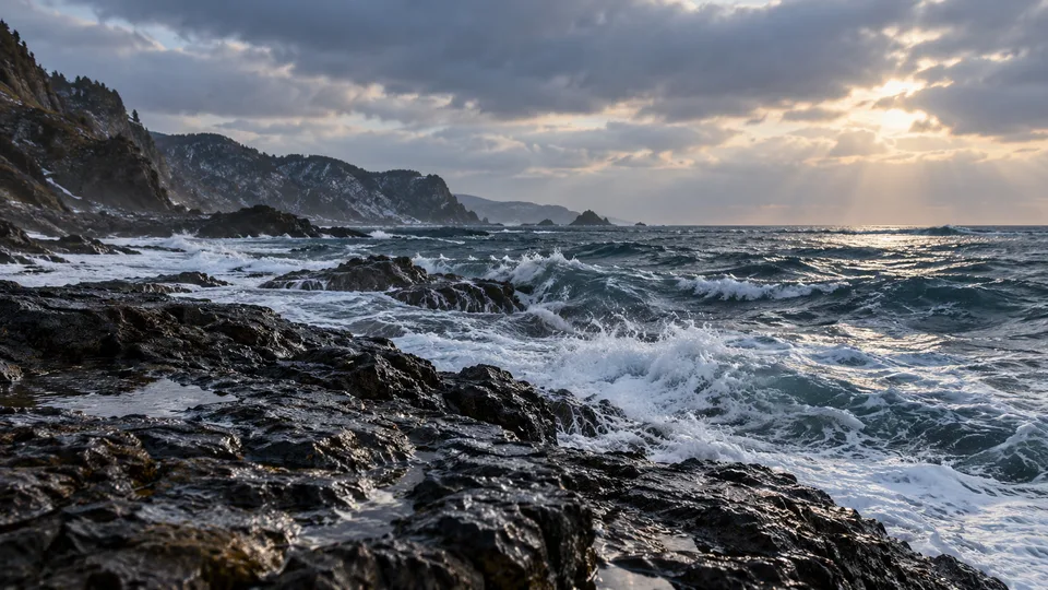 Cold blue-gray waves break against an anonymous rocky Japanese shoreline under broken winter cloud.