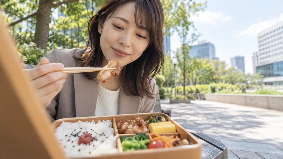 A young Japanese woman picks up a bite from a compartmented bento on a quiet outdoor bench.