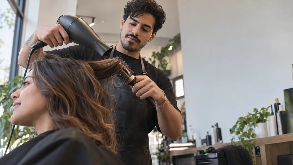 A hairdresser works with a brush and dryer while a client sits in a small salon.