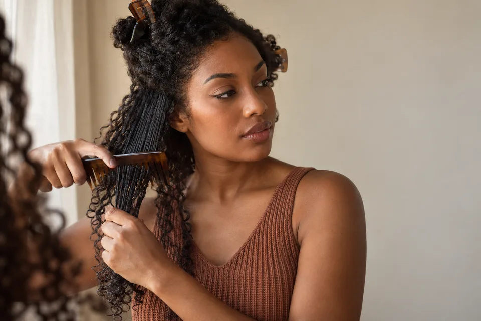 A woman separates and gently combs damp curls beside a softly lit window.