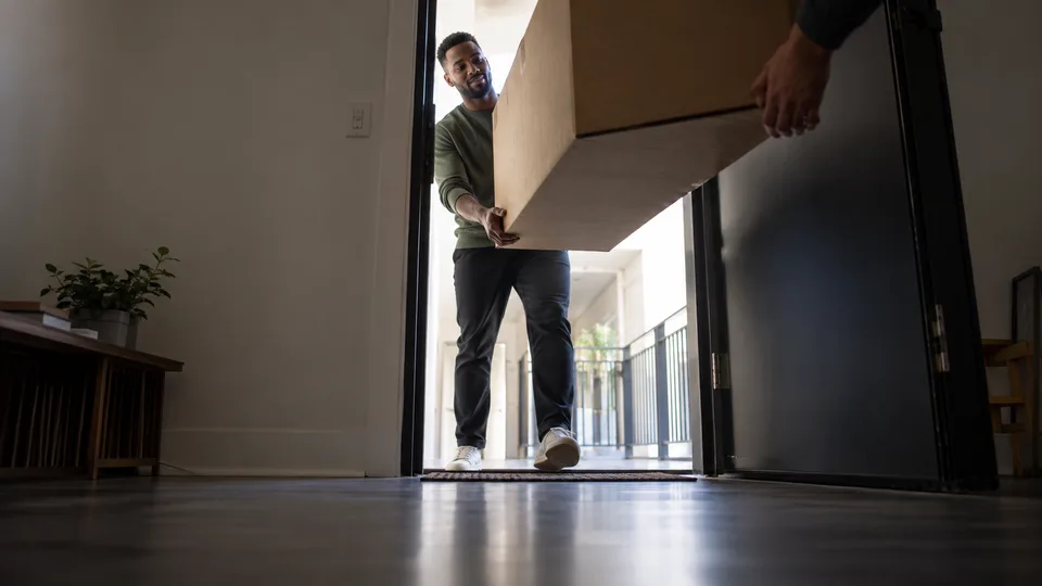 A new resident guides a plain moving box through an apartment doorway from the bright corridor outside.