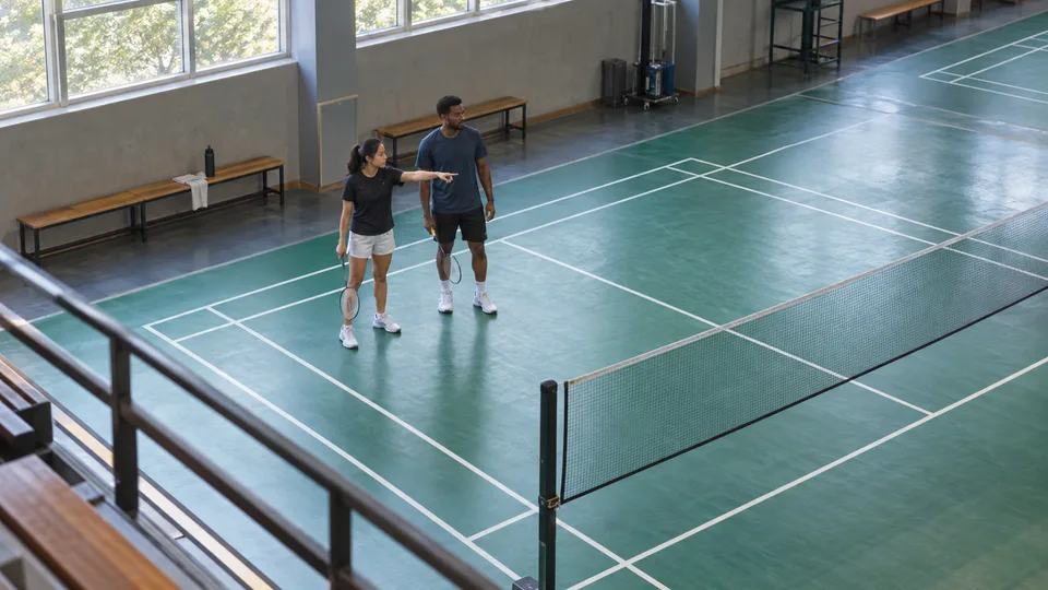 Two badminton partners point and confer on their side of an indoor court before the next rally.